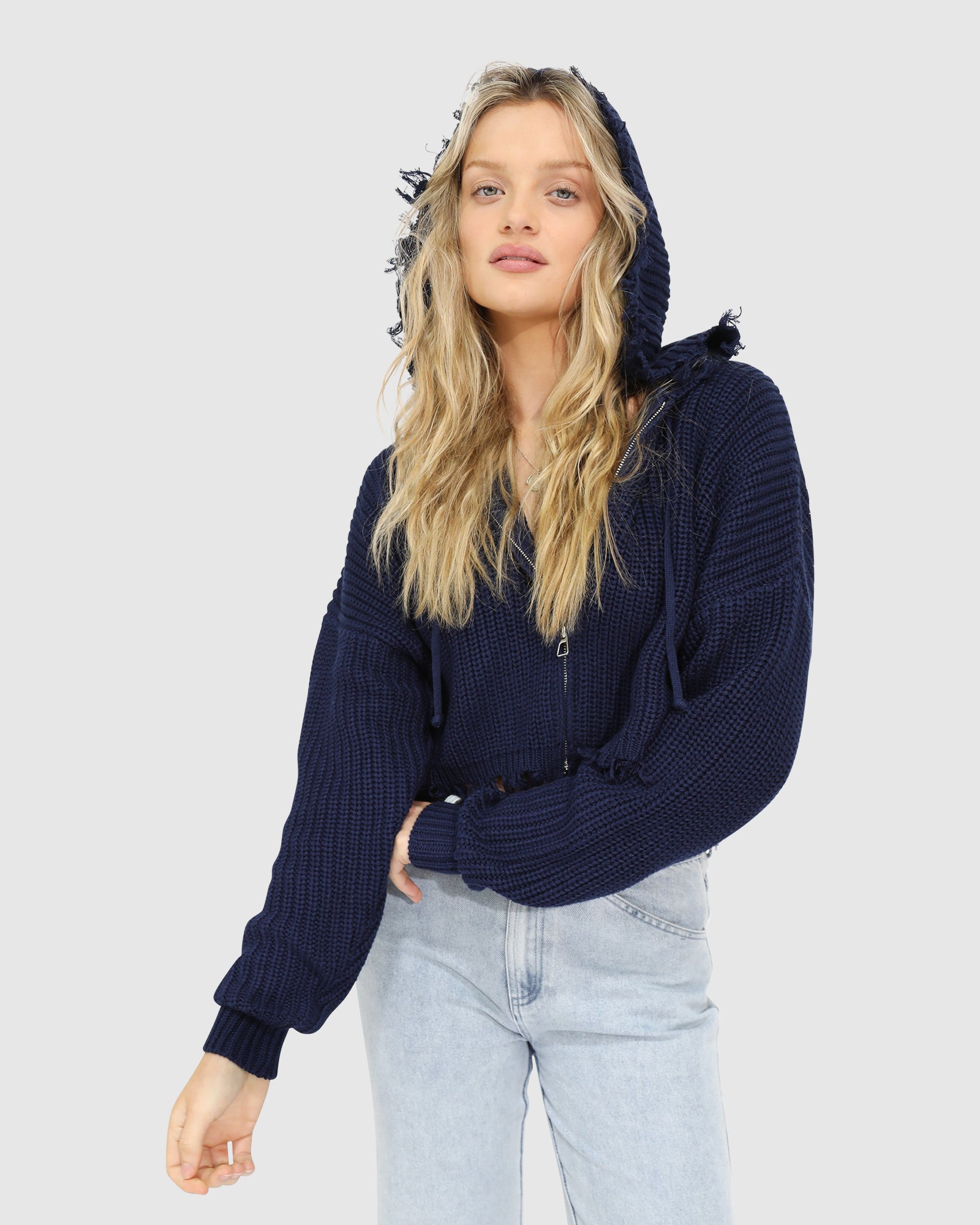 Woman wearing the Another Love Fringe Knit Zip Cardigan in navy, featuring chunky knit texture, zip front, drawstring hood, and fringed cropped hem – Belle and Bloom knitwear.