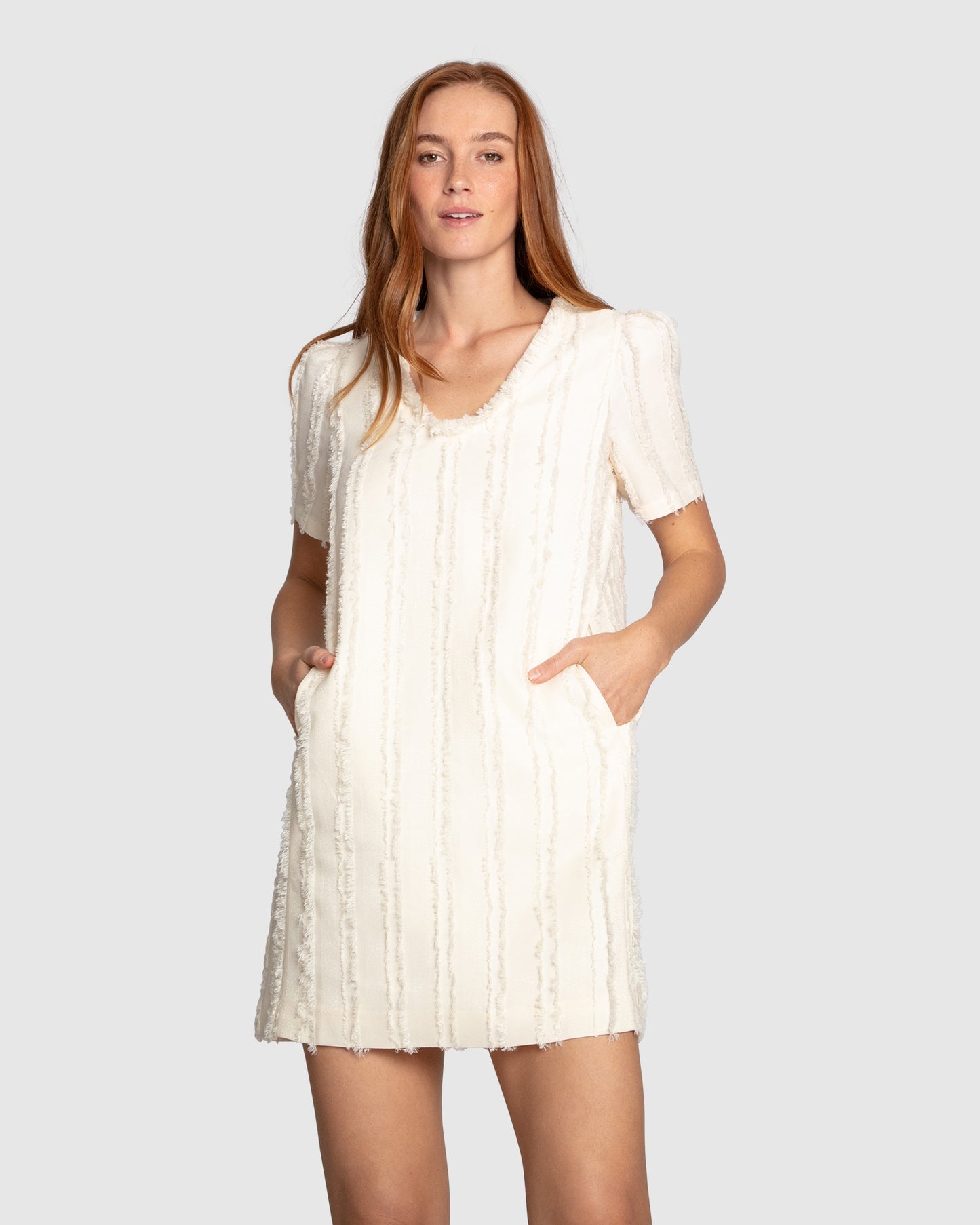 Woman wearing Belle & Bloom's cream-coloured Solana Boho Fringe Mini Shift Dress with a frayed V-neckline, textured sleeveless shift silhouette, side-seam pockets, and subtle fringe detail at the hem.