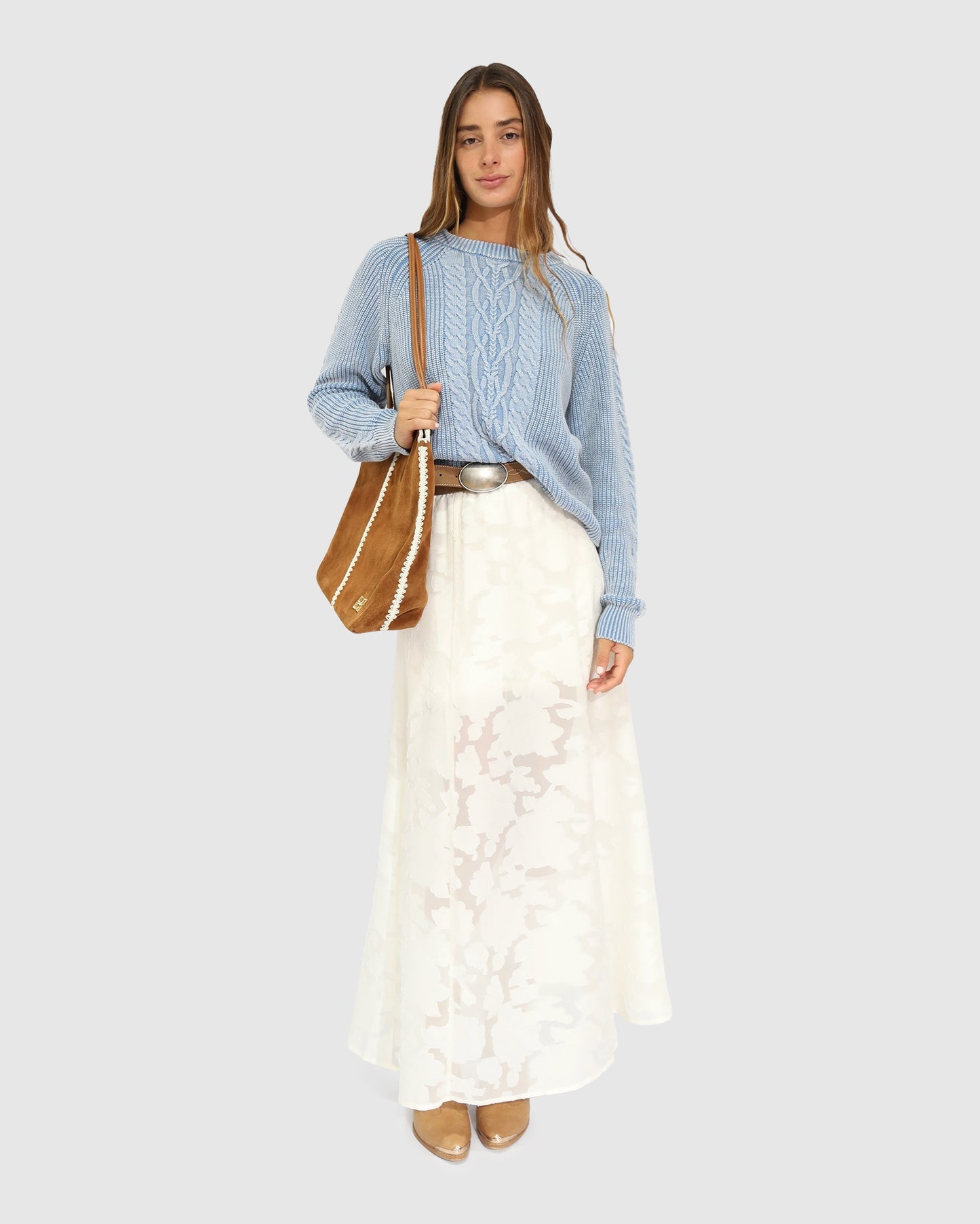 Woman wearing a blue sweater and white lace skirt with a brown bag on a light gray background