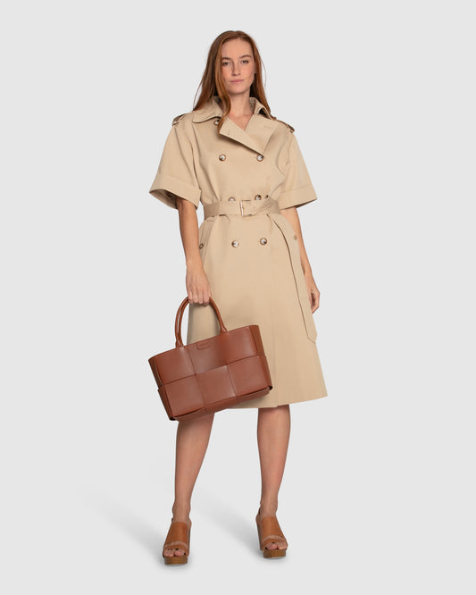 Woman wearing Belle & Bloom Dust & Daring Short Sleeve Trench Style Dress in beige with belted waist, double-breasted buttons, tan tote bag and platform sandals.