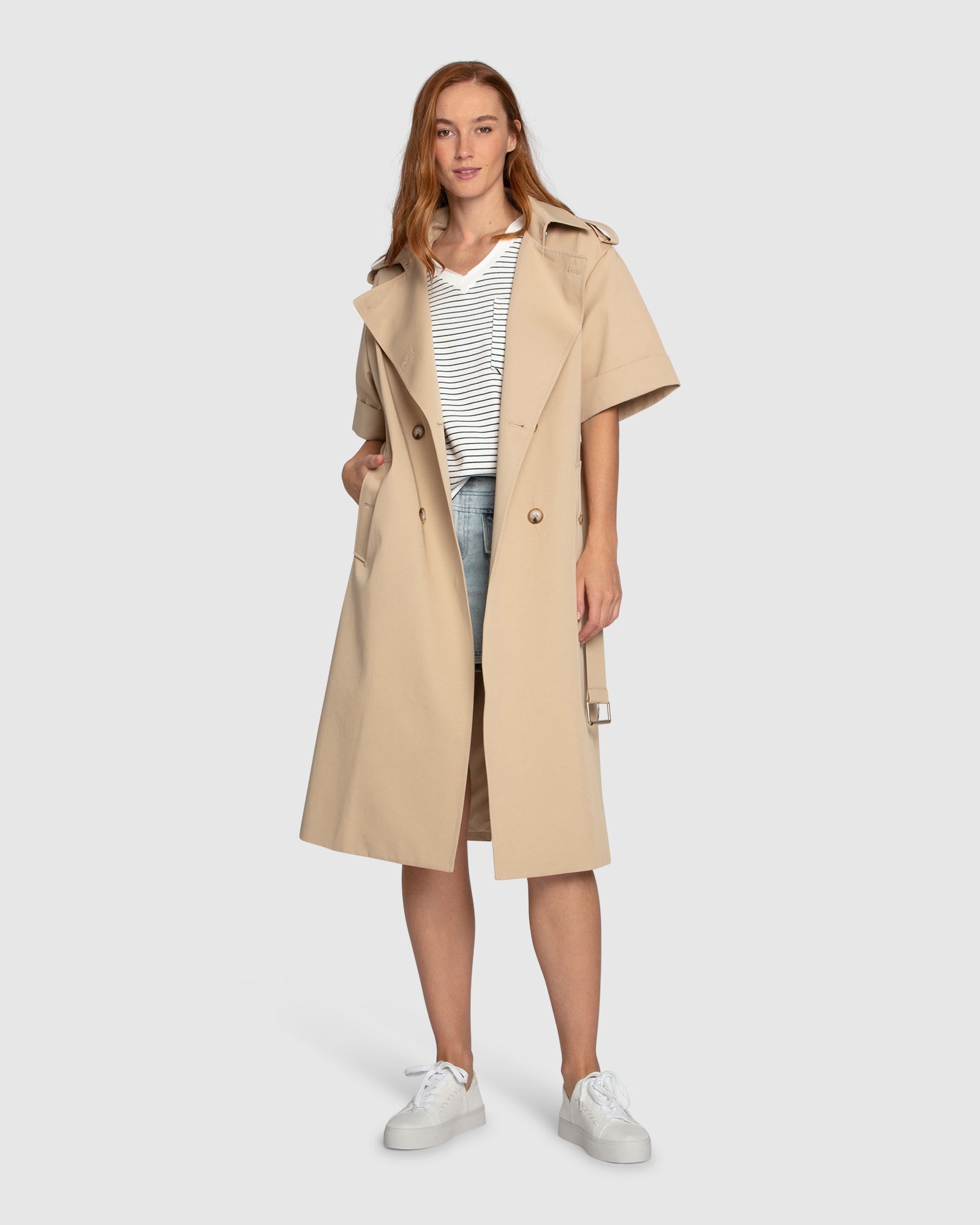 Woman wearing Belle & Bloom Dust & Daring Short Sleeve Trench Style Dress in beige with belted waist, double-breasted buttons, tan tote bag and platform sandals.