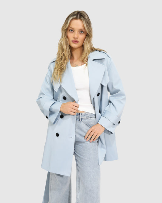 Woman wearing the Whispers of Verona Trench Coat in sky blue, featuring a longline double-breasted silhouette, belted waist, storm flap, and classic lapels – Belle and Bloom outerwear.