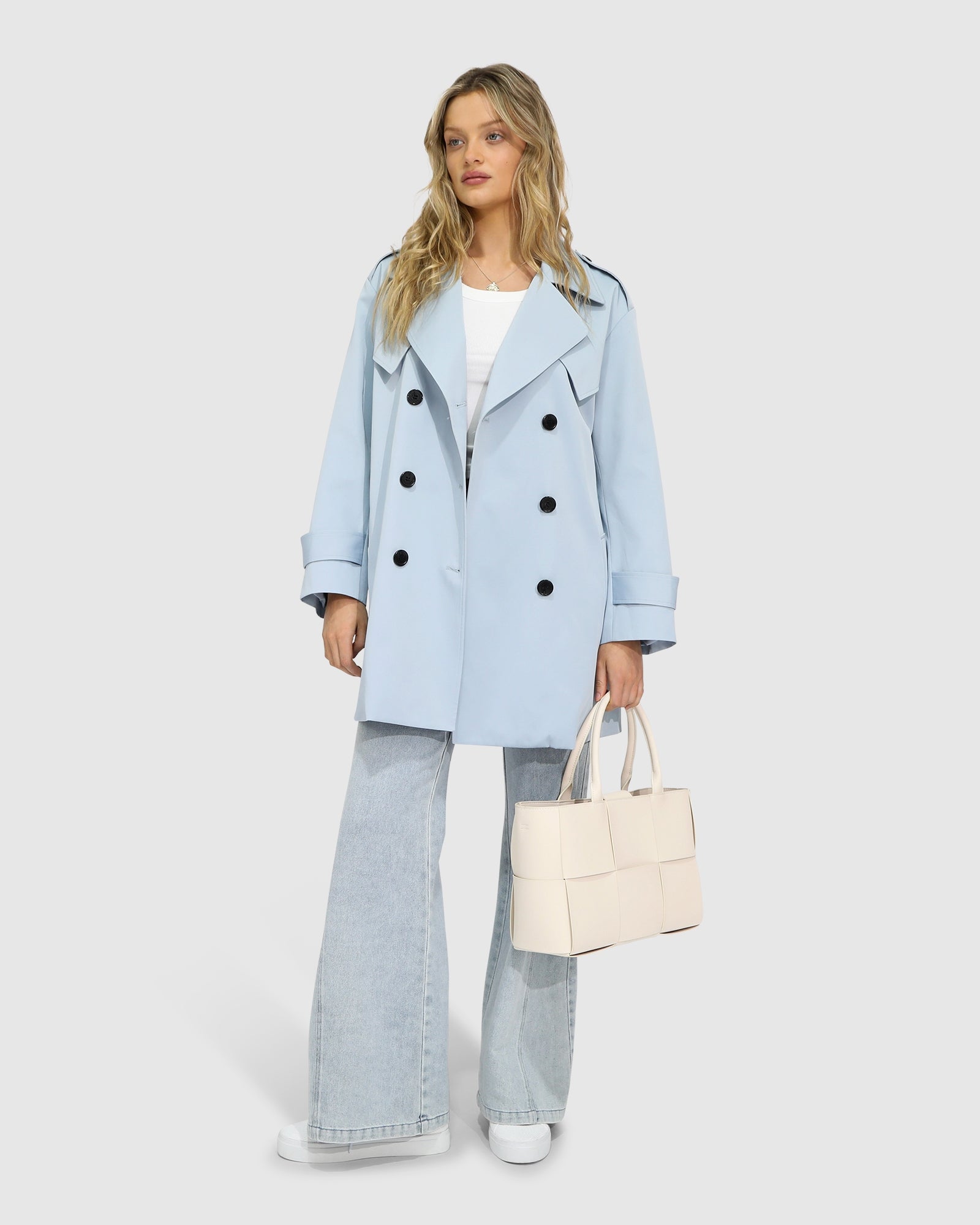 Woman wearing the Whispers of Verona Trench Coat in sky blue, featuring a longline double-breasted silhouette, belted waist, storm flap, and classic lapels – Belle and Bloom outerwear.