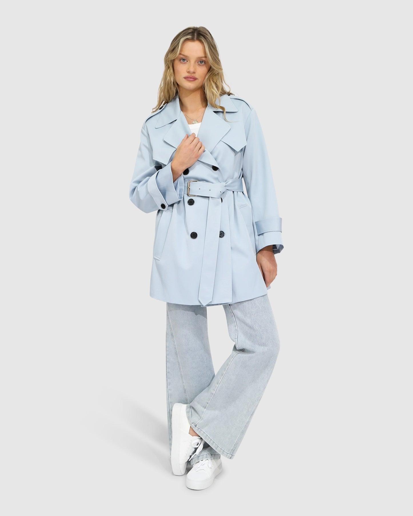 Woman wearing the Whispers of Verona Trench Coat in sky blue, featuring a longline double-breasted silhouette, belted waist, storm flap, and classic lapels – Belle and Bloom outerwear.